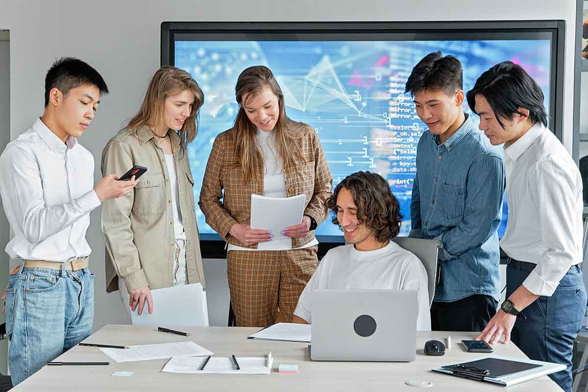 A group of six men and women standing around an office table and looking at a laptop and papers.