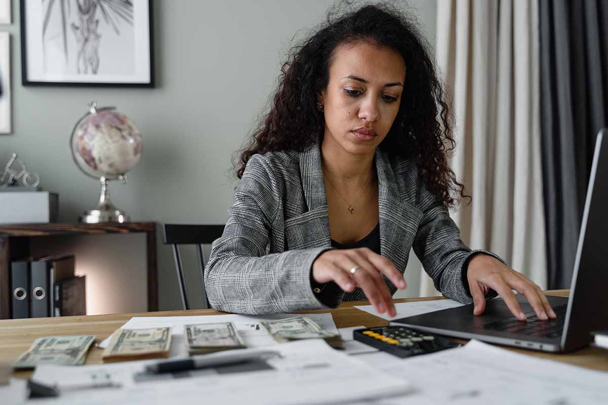 Business woman sitting at an office desk typing on a laptop and using a calculator.