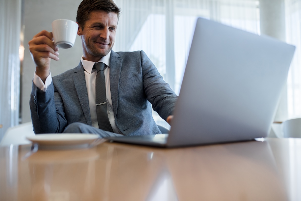 Happy young businessman using laptop and having coffee at office. Male executive working on laptop during coffee break.