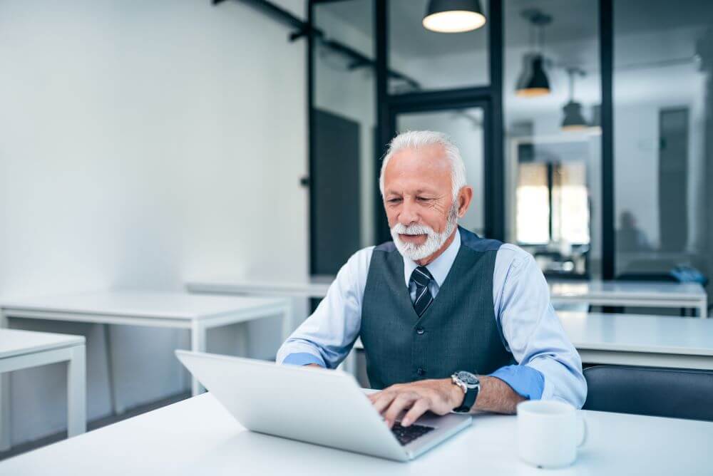 senior man smiling and working on a laptop in an office.