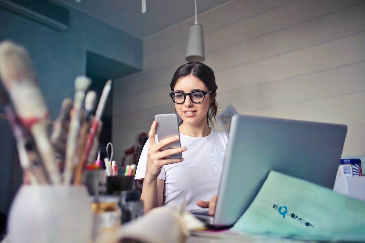 Woman with glasses works on laptop while looking at smartphone