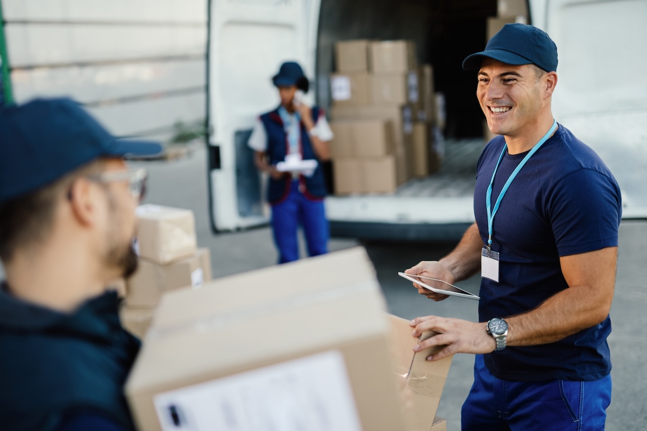 Happy manual worker using touchpad while communicating with his coworker and organizing package delivery.