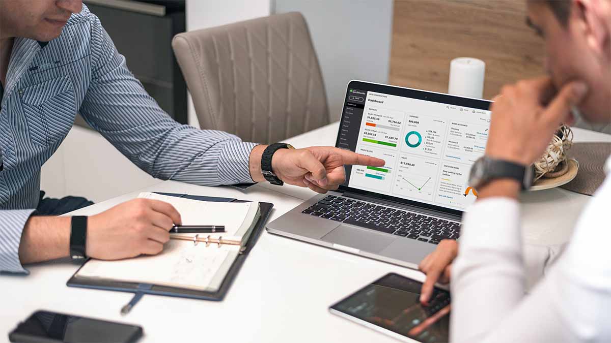 Two men sitting at an office table. One of the men is pointing at a laptop screen displaying the QuickBooks Online dashboard.