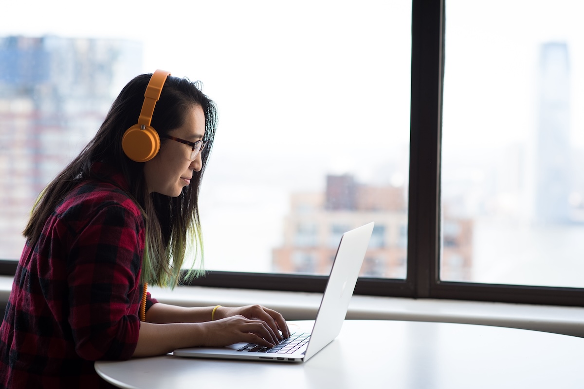 Woman with orange headphones works on laptop in front of window