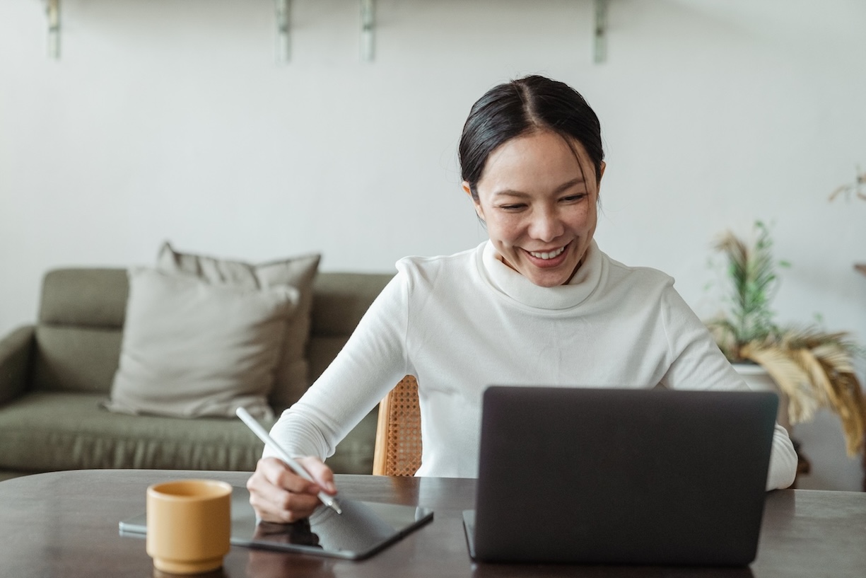 Smiling woman looking at laptop screen while taking notes on notebook