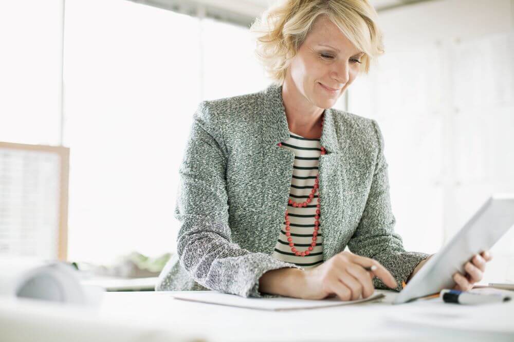 Senior lady with a grey blazer clicking on a tablet screen and smiling.