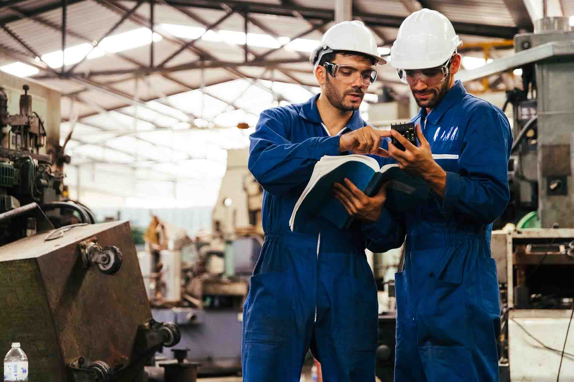 Two workers wearing hardhats and blue coveralls. One is holding a book and they are looking at a machine part.