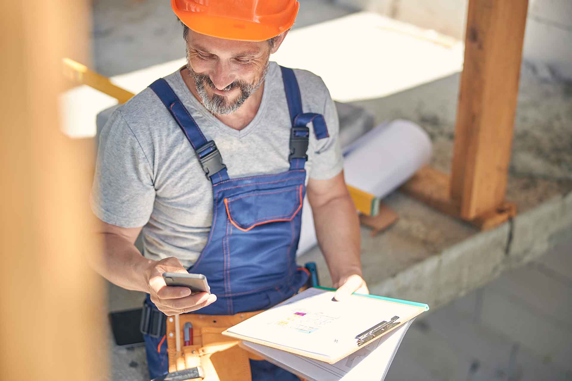 A man wearing a hardhat and overalls holding a clipboard on a construction site.