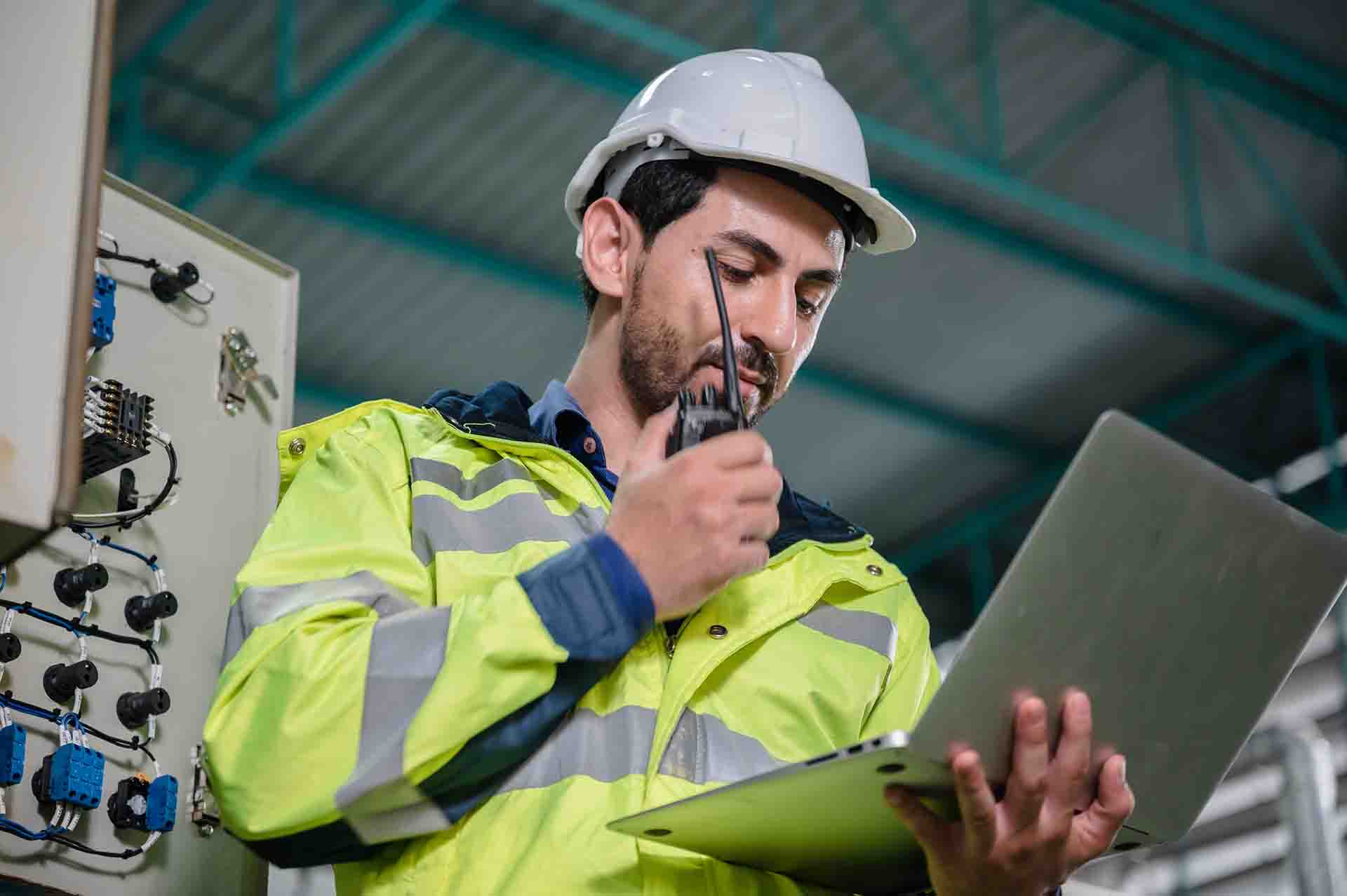A man wearing a hardhat and high visibility jacket looking at a laptop and speaking on a walkie talkie.