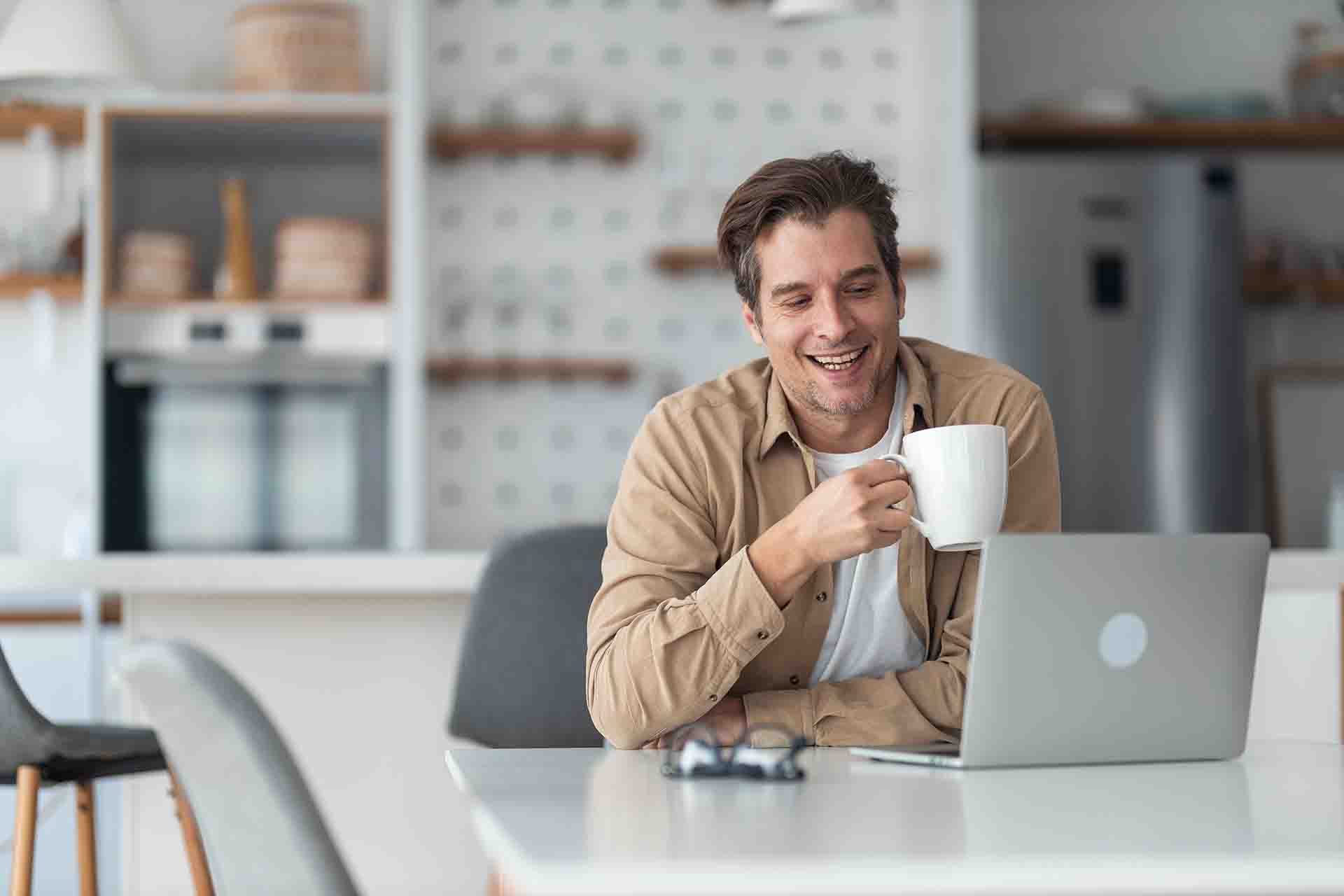 A man sitting a table with a laptop and holding a mug.