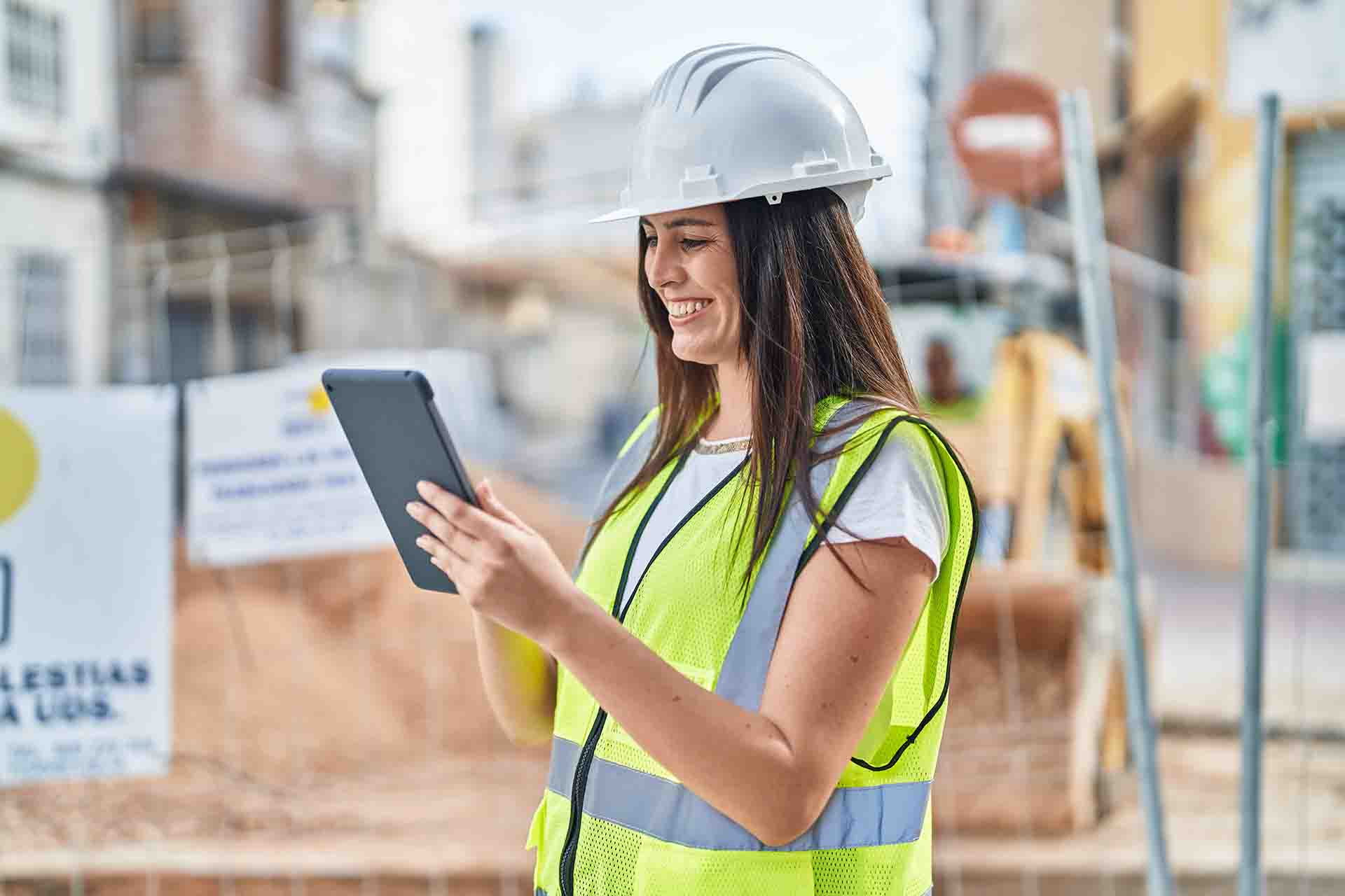 A woman wearing a hardhat and high visibility vest looking at a tablet while standing near street construction.
