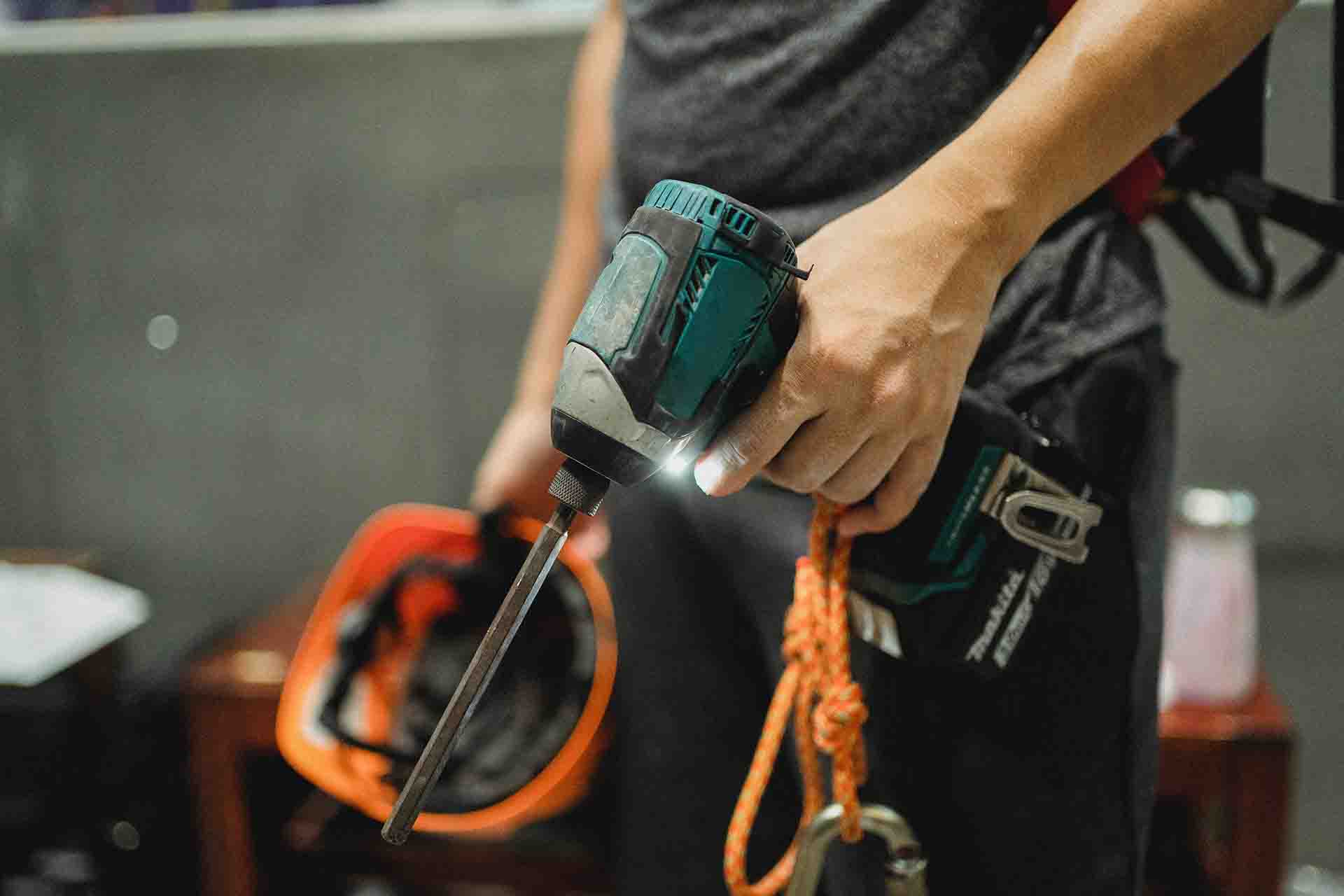 A cropped photo showing a person holding a screwdriver in one hand and a hard hat in the other hand, and also wearing a utility belt.