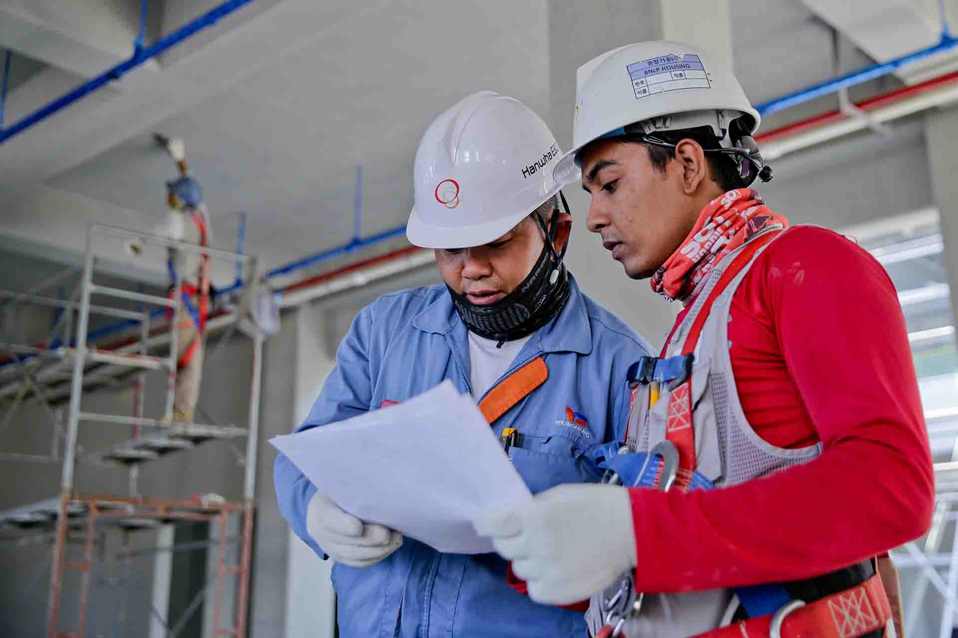 Two men wearing overalls and looking at a document together while standing inside a construction site.