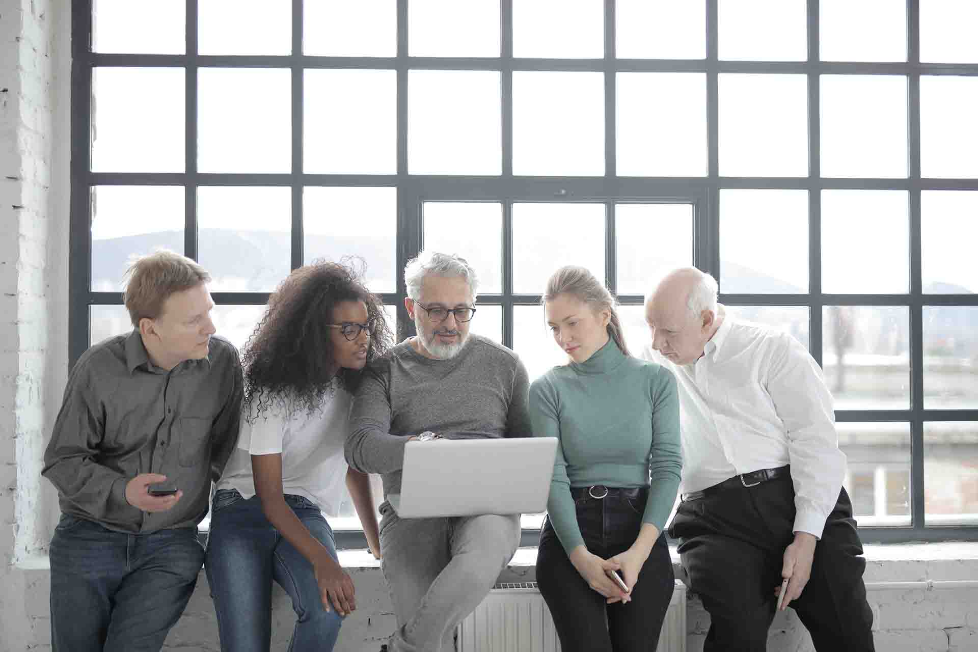 A group of five people sitting in a row while looking at a laptop together.