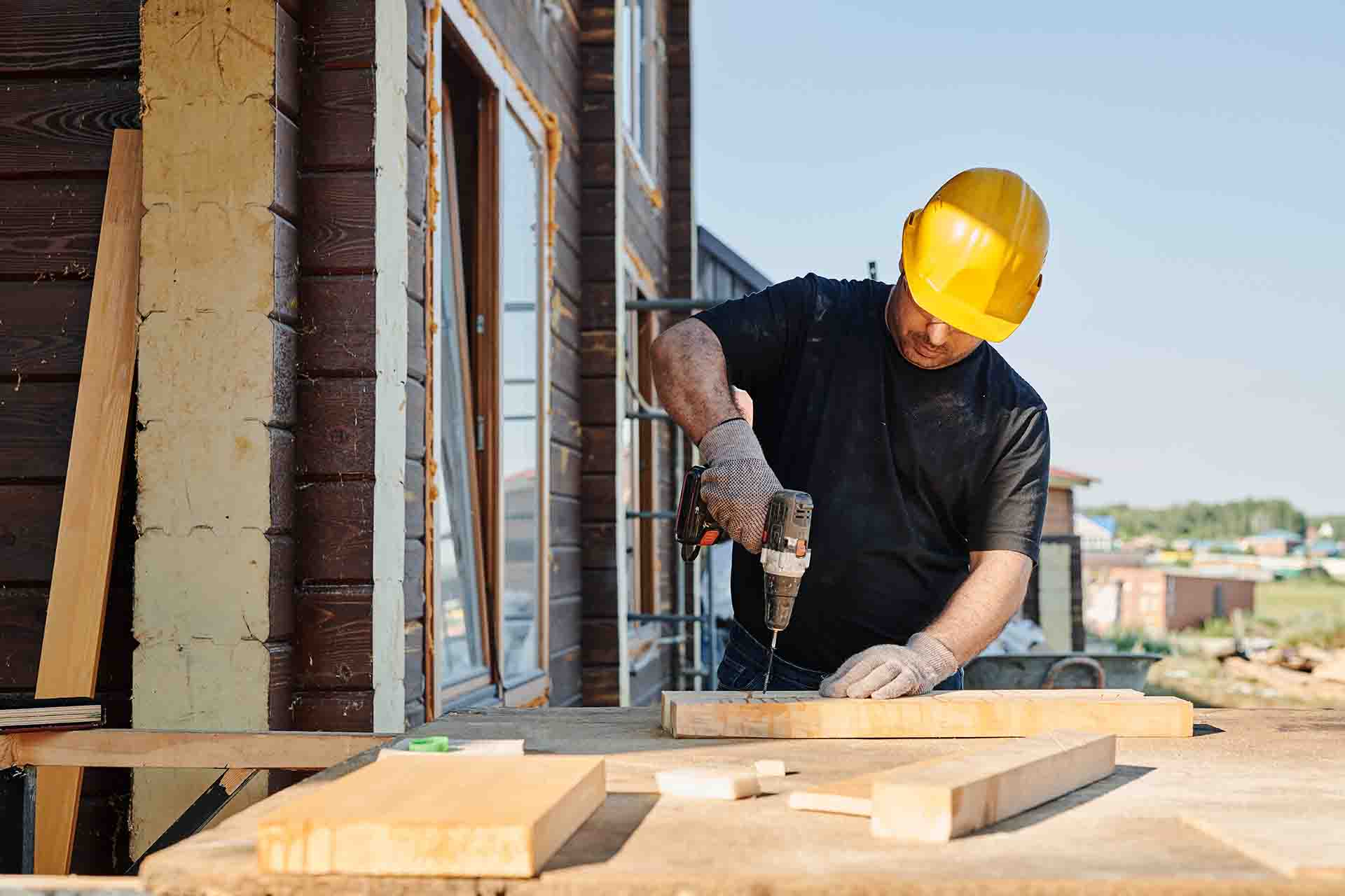 A man wearing a construction hat and using a power drill on wood parts outside of a home under construction.