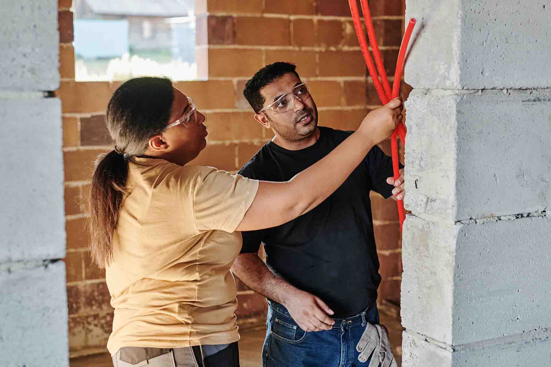 A man and a woman wearing safety glasses and holding orange tubing on a construction site.