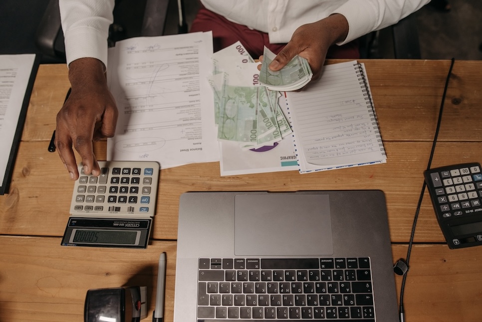 Man Sitting at the Desk and Counting Money while Using a Calculator