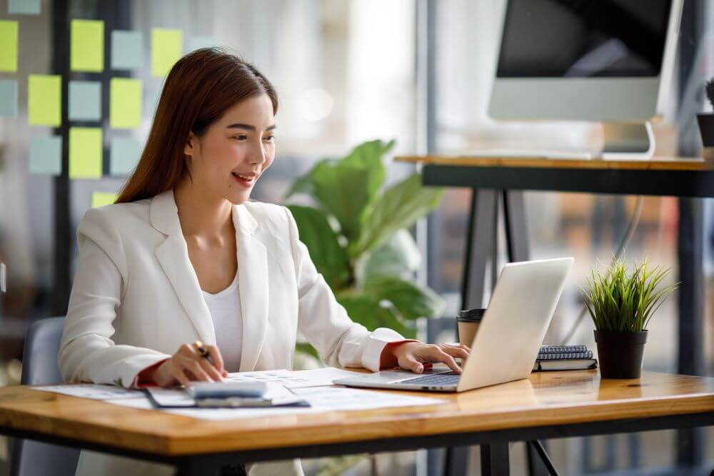 young woman in a white blazer smiling while working on a laptop