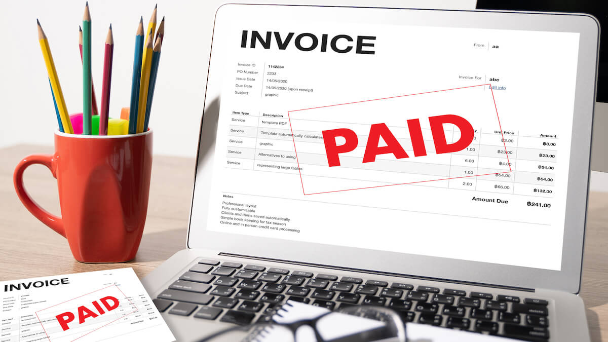A laptop screen showing a paid invoice, with said invoice sitting on the desk.